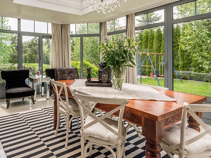 Sunroom surrounded by large windows looking out to the green garden with large rectangular wooden table and white wooden chairs.